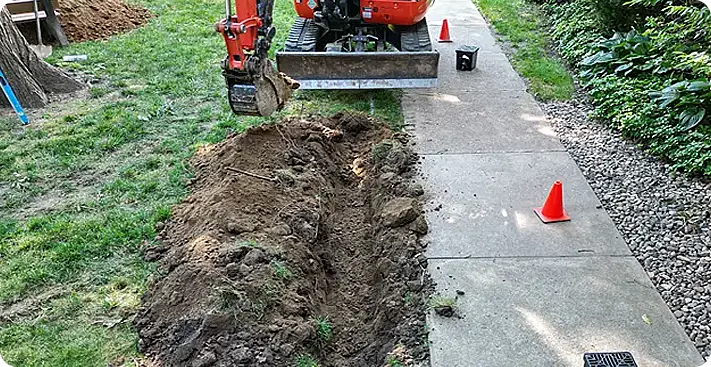 Trench excavation next to a concrete walkway in Media, PA, with compact utility excavator and safety cones part of a drainage & grading solution for residential landscape improvement.