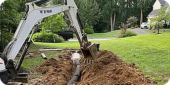 Excavator backfilling a drainage trench with newly installed pipe in a residential yard.