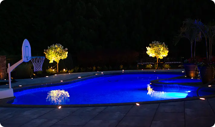 Poolside patio with LED lighting and illuminated landscape trees at night. All products of ScapeWorx superior craftsmanship.