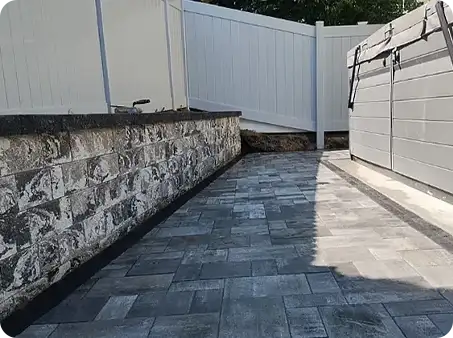 Paver walkway next to a stone retaining wall with a white fence.