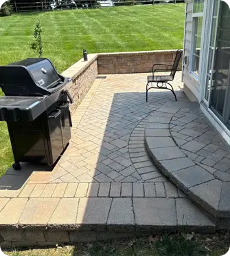 Landscaping in Kennett Square with paver patio designed by ScapeWorx featuring a curved step, built-in retaining wall seating, and grill setup on a sunlit outdoor space.